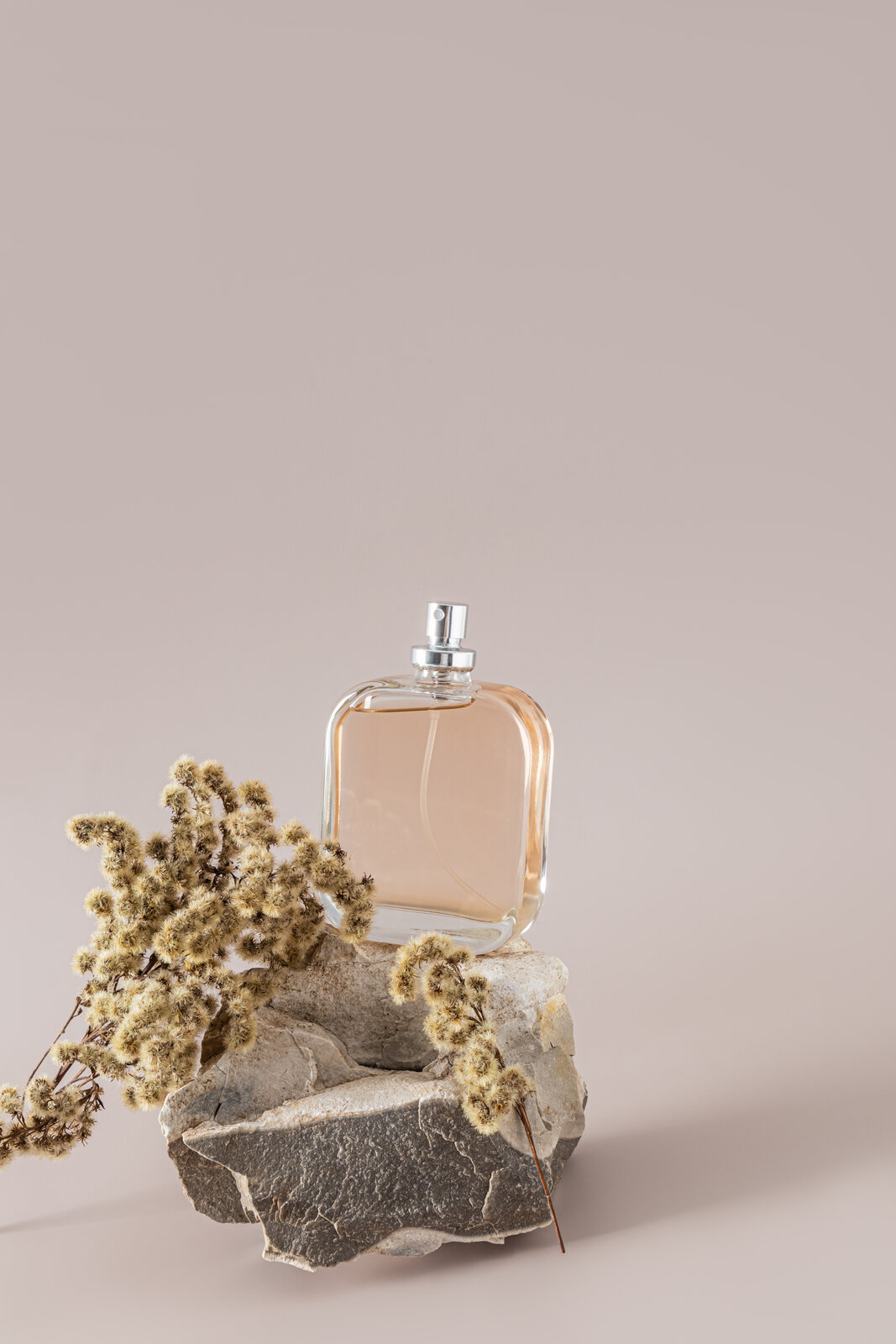 A perfume bottle with a silver cap resting on a natural stone, styled with dried botanical branches against a warm nude backdrop